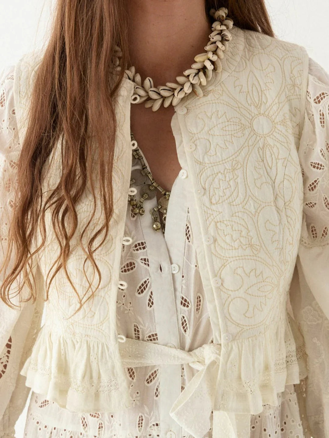 A person wearing a white embroidered blouse and the Sandro Vest - Tesela Ivory from Maison Hotel. They accessorize with a necklace featuring shell-like elements and a cross pendant.