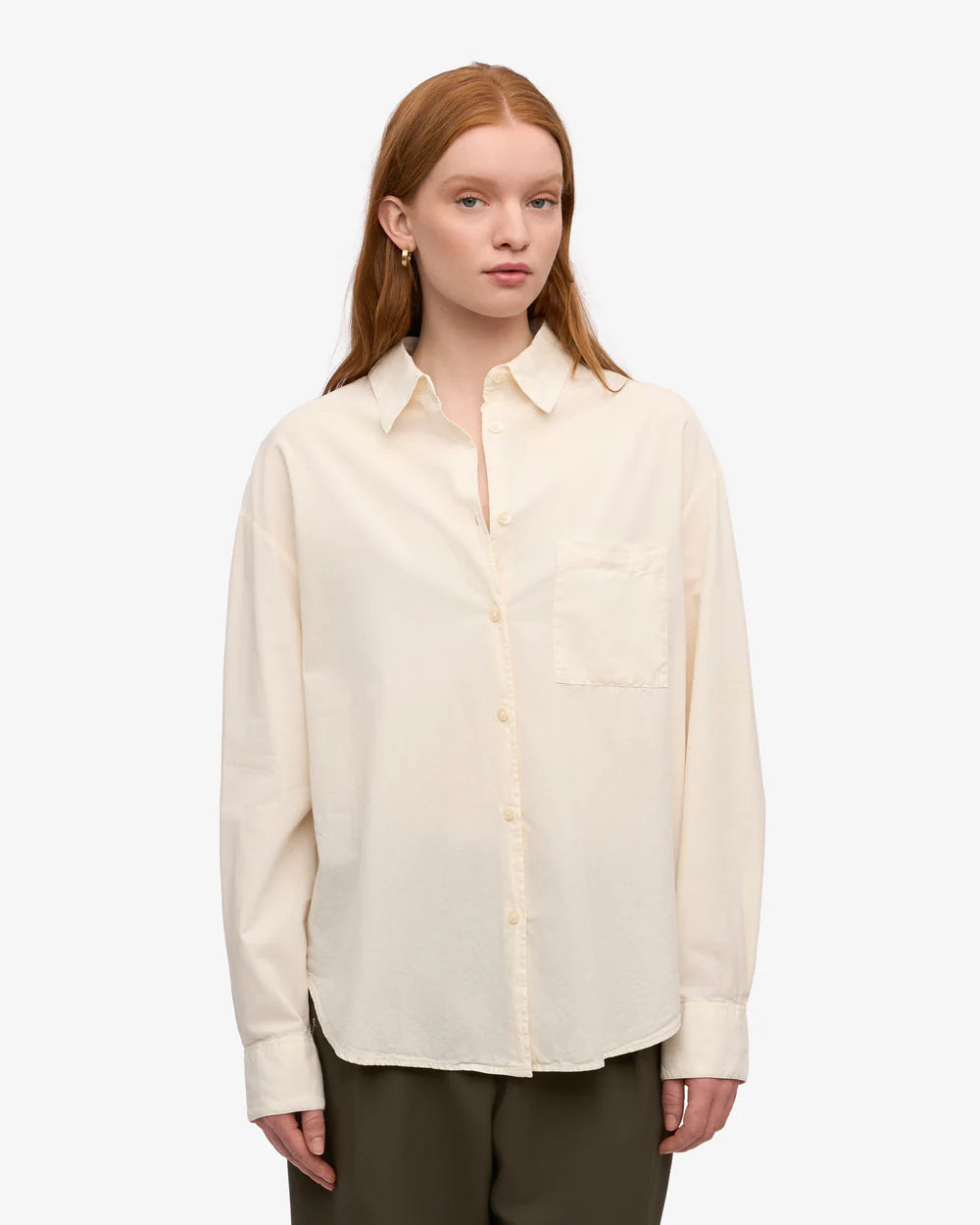 A person with long reddish hair wears the Colorful Standard Organic Oversized Shirt in cream, featuring a chest pocket, paired with dark pants and standing against a plain white background.