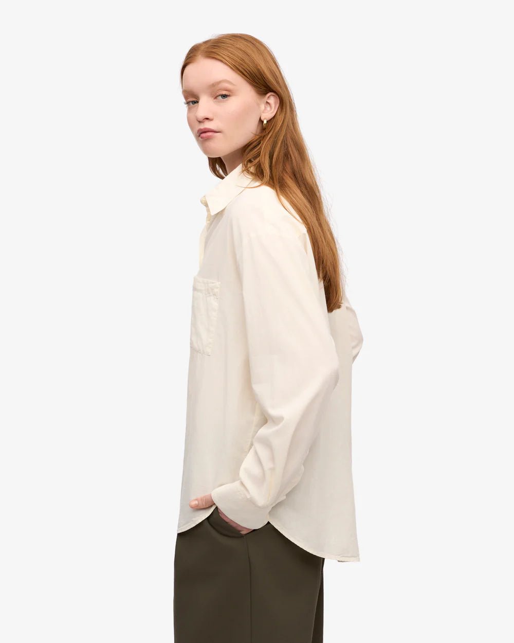 A woman with long red hair models the Colorful Standard Organic Oversized Shirt in a creamy shade, paired with dark pants and standing casually with one hand in her pocket against a plain background.
