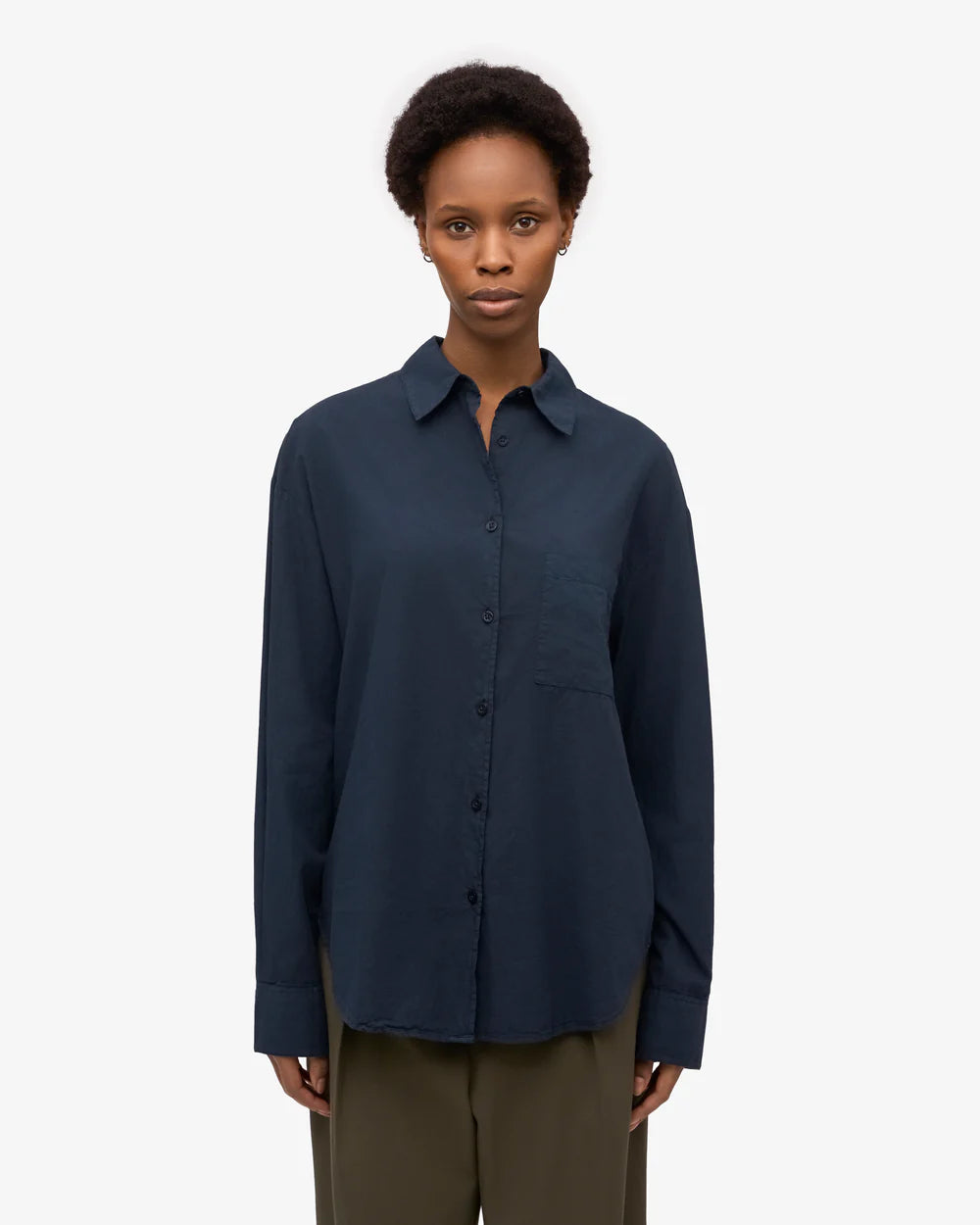 A person stands facing forward against a plain white background, wearing the Colorful Standard Organic Oversized Shirt in dark blue with a chest pocket, paired with olive green pants.