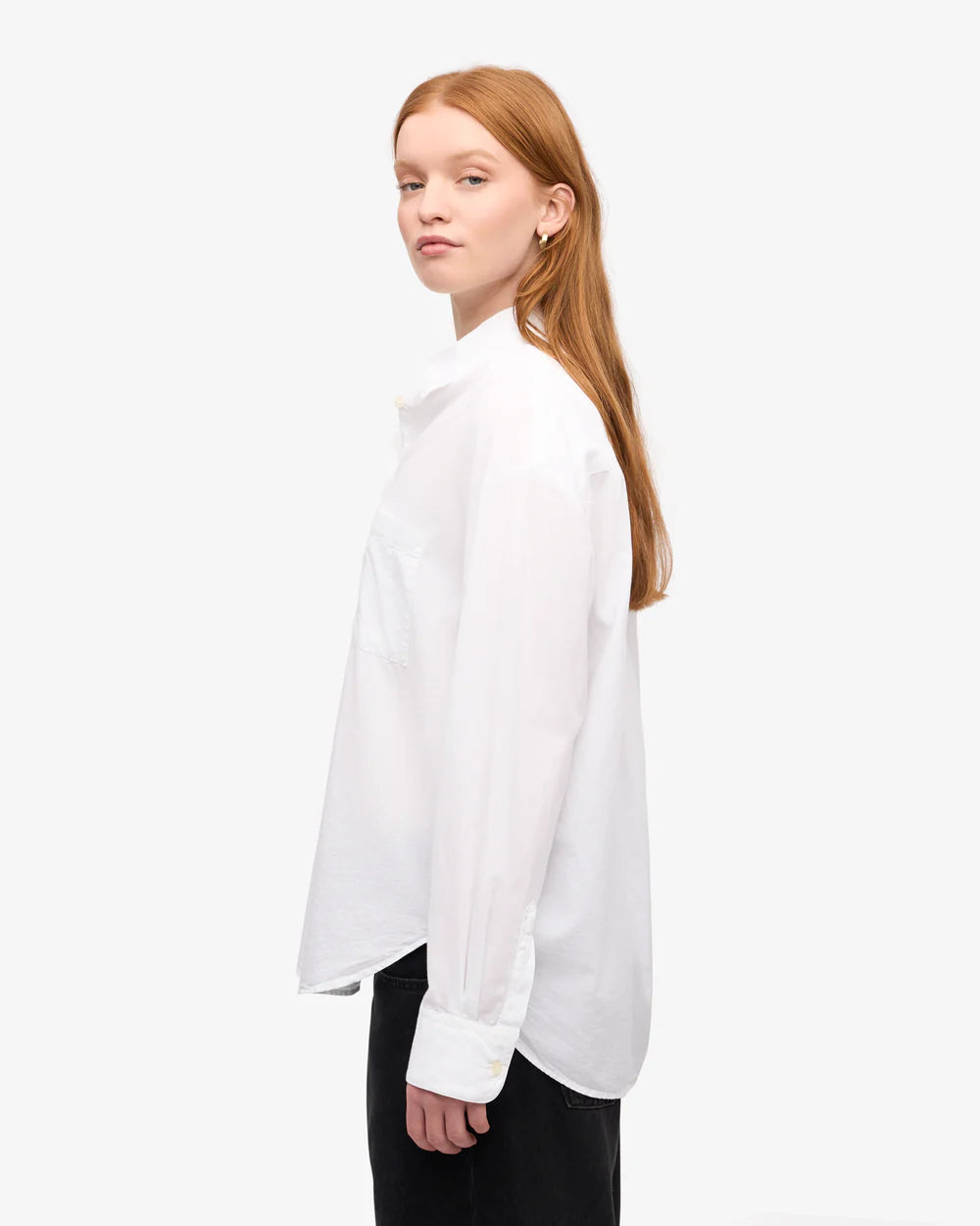 A person with long reddish hair, facing sideways against a plain background, wears the Organic Oversized Shirt by Colorful Standard paired with black pants.