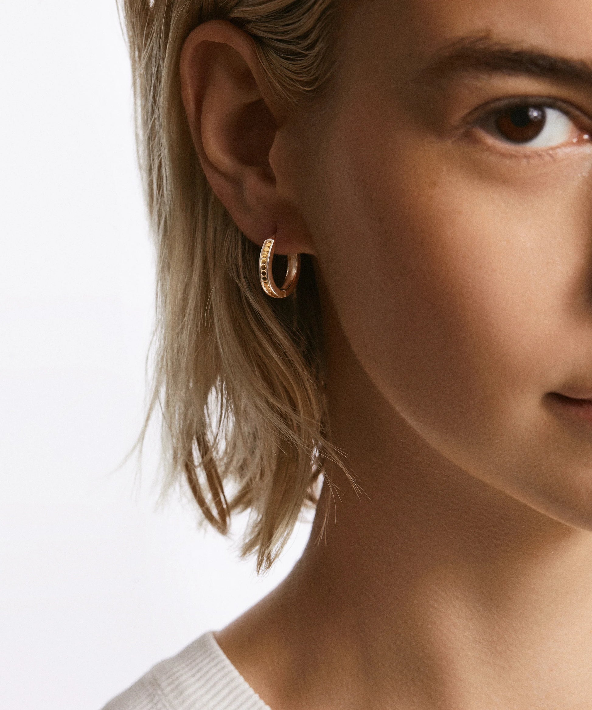 Close-up of a person with light hair wearing Anna Beck's Classic Small Hoop Earrings in 18k gold plating and a white top, showing one side of their face and ear.