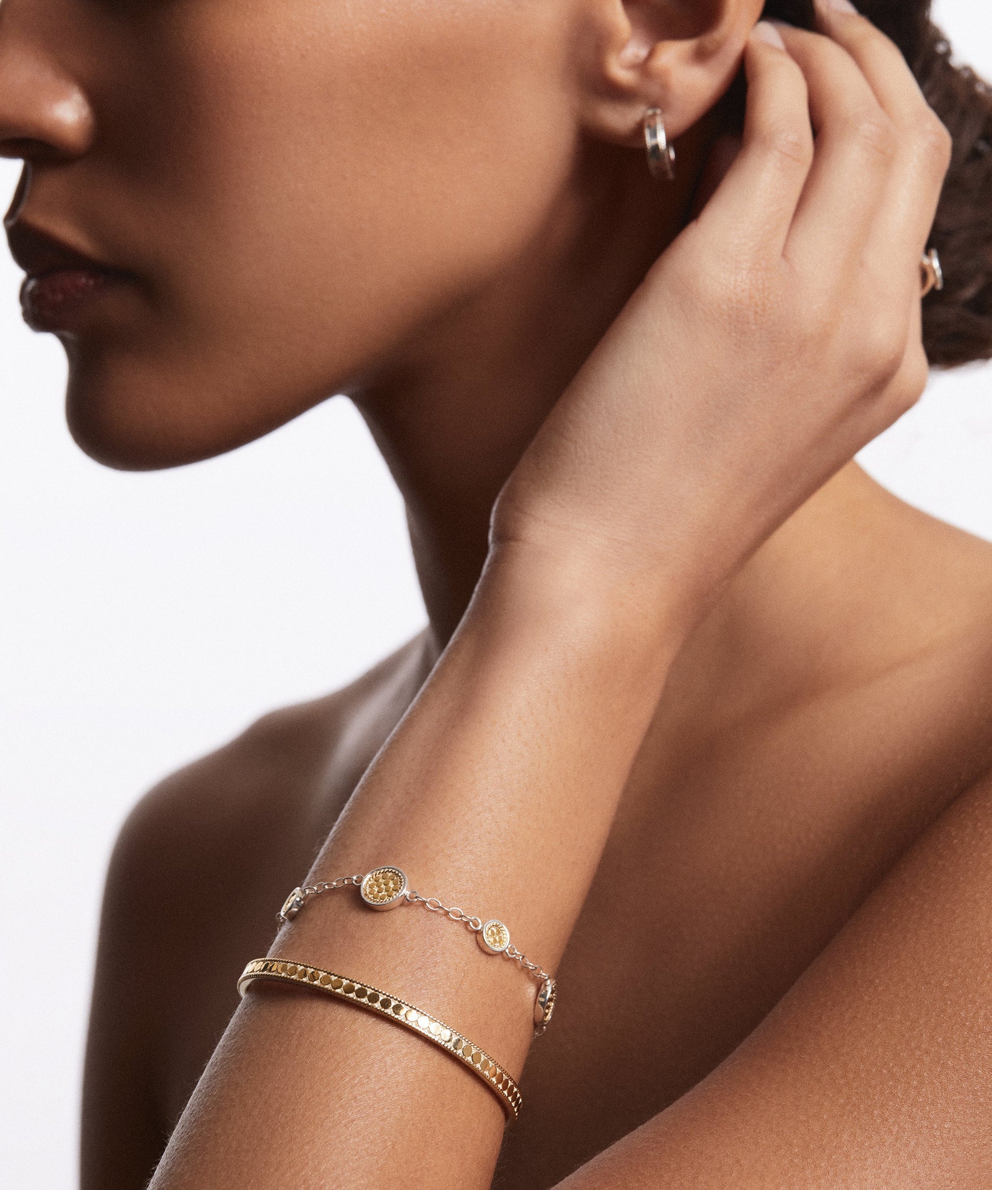 A person with brown skin touches their ear, showcasing Anna Beck's gold hoop earrings and a gold bracelet alongside the Classic Stacking Cuff - Gold. The jewelry highlights intricate Balinese techniques and 18k gold plating against a white background.