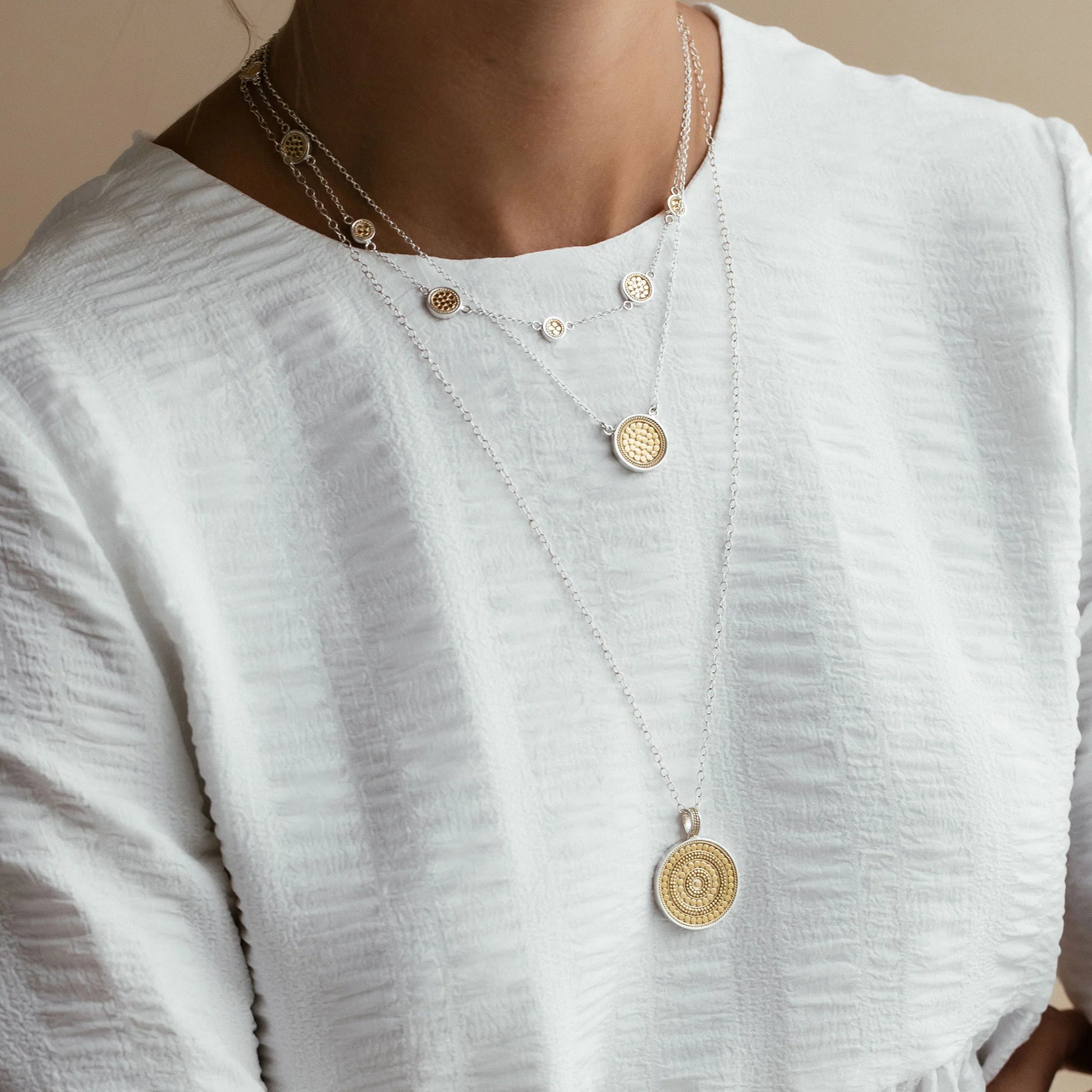 A person wearing a textured white blouse and the Anna Beck Classic Station Necklace - Gold & Silver, featuring handcrafted layered medallions with 18k gold plating.