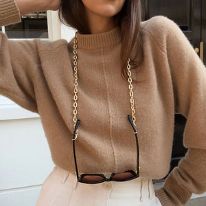 Gold sunglasses chain by Talis Chains styled by a person wearing a chunky beige knitted jumper and nude pants, stanfing infront of a white wall and brown wooden door to the right. 