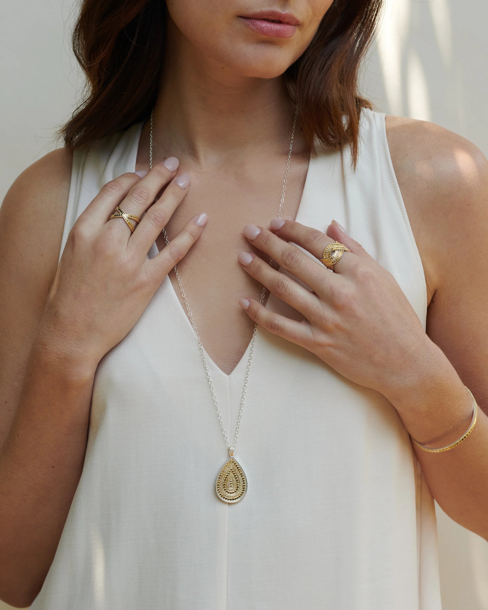 A woman wearing a sleeveless white top and several gold rings displays the Anna Beck Classic Large Teardrop Necklace, which is handcrafted and reversible, featuring a pendant on a sterling silver chain and bracelet.