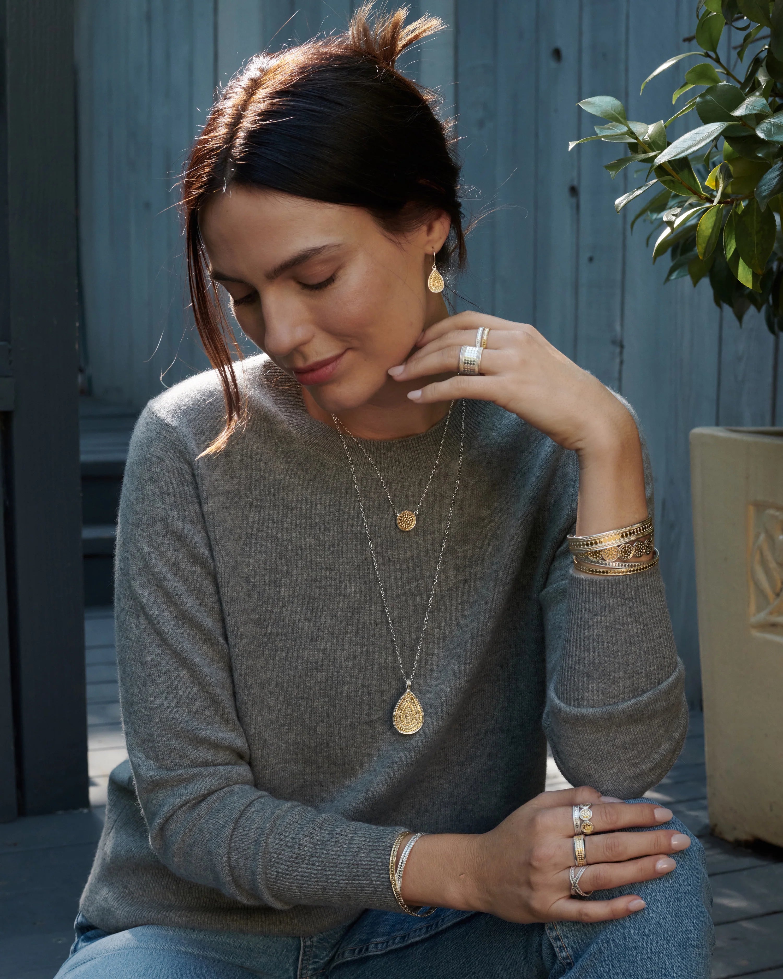 A woman with dark hair in a loose bun sits, wearing a gray sweater and various gold jewelry including rings, bracelets, and Anna Beck's Classic Large Teardrop Necklace - Reversible on a sterling silver chain. She is looking down with one hand resting on her chin.