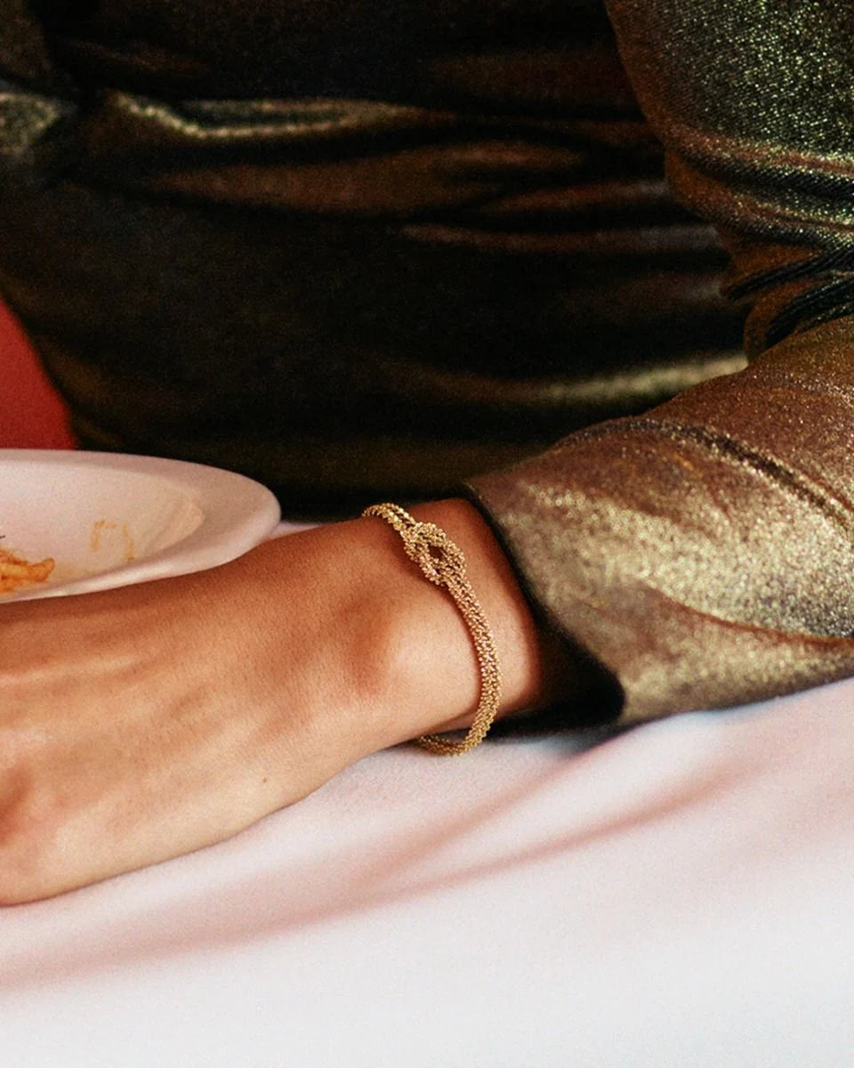 Close-up of a person wearing the Tinsel Chain Bracelet by EDBLAD, gleaming with festive sparkle on their wrist above a green sleeve. It rests elegantly on a white table near a plate.