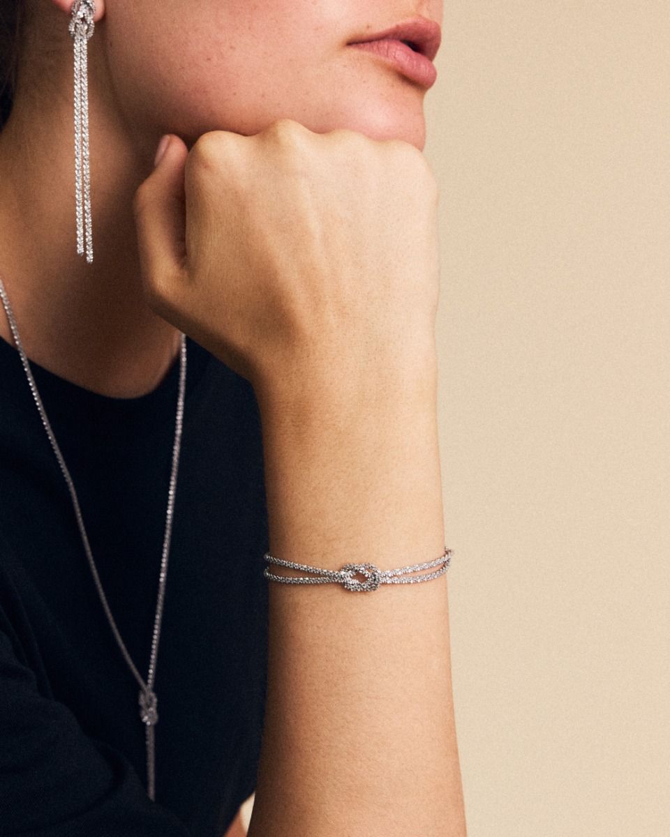 Person adorned with silver jewelry: earrings, a necklace, and the EDBLAD Tinsel Chain Bracelet featuring a knot design and festive sparkle, resting chin on hand.