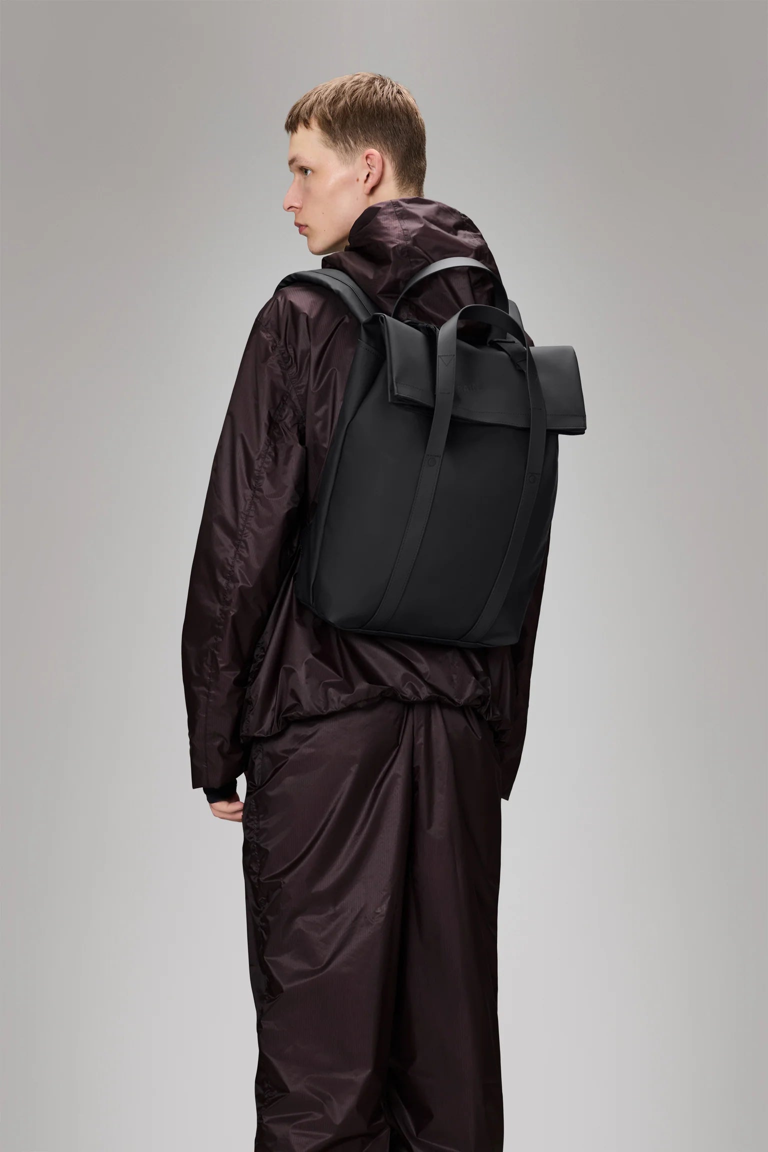 A person wearing a dark, shiny jacket and pants set is seen from the side and back, carrying the Rains 2 Way Tote Backpack in Black against a plain gray background.