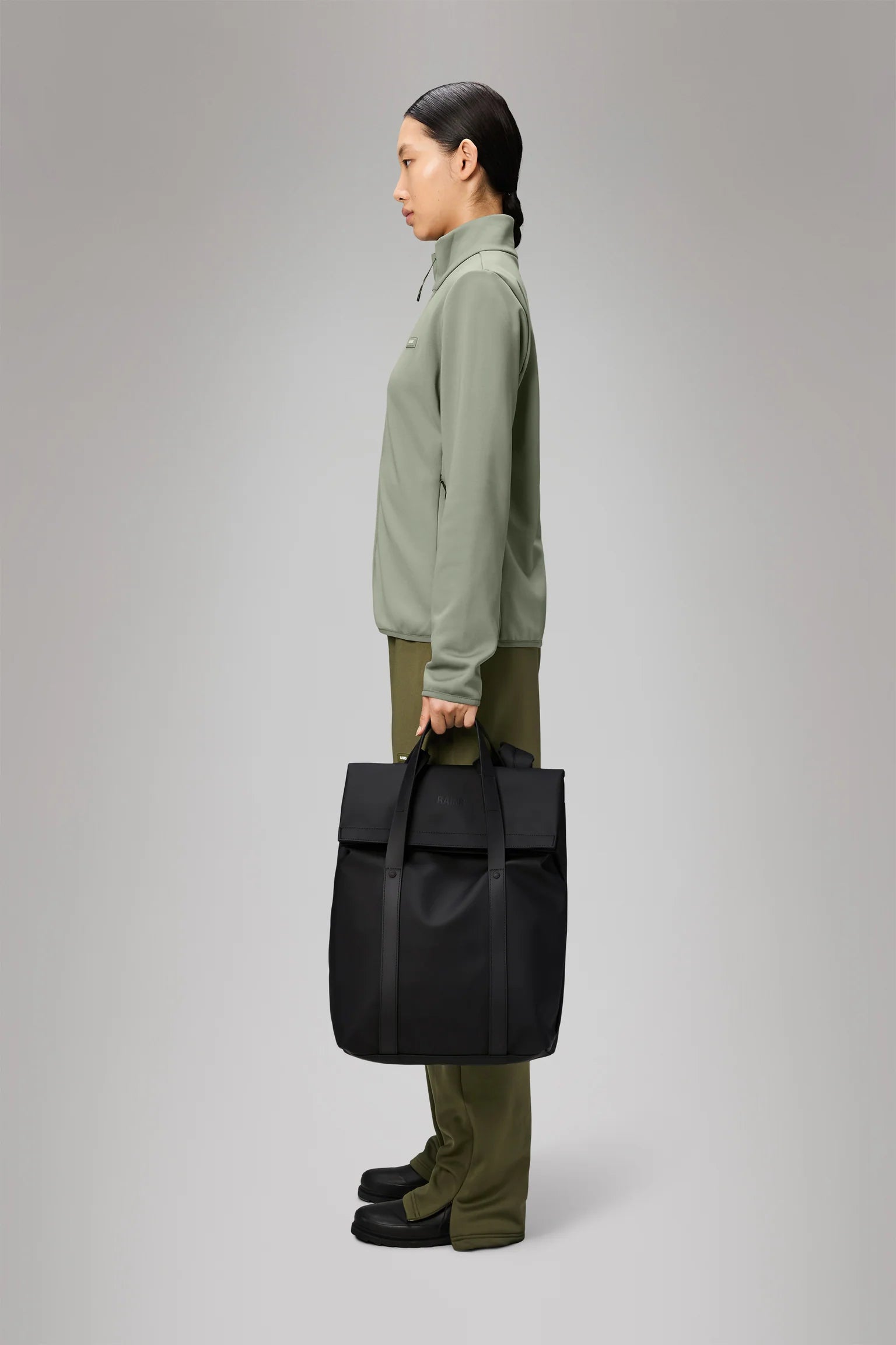 A woman in profile holds the Rains 2 Way Tote Backpack in black, dressed in a light green zip-up jacket, olive pants, and black shoes against a plain gray background.
