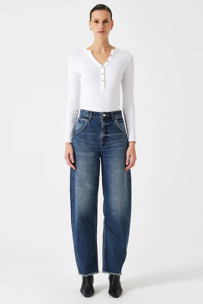 A person faces forward against a plain white background, wearing Seventy & Mochi's Bo Barrel Leg Jeans in Phoenix, a white long-sleeve henley top, and black shoes.