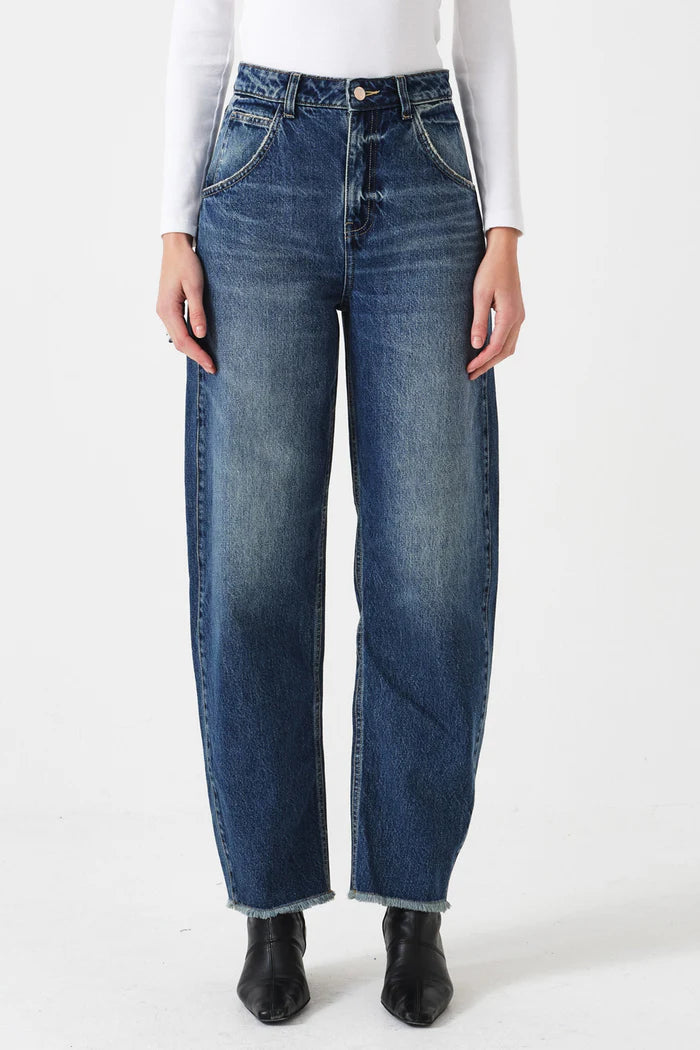 A person wears Seventy & Mochi's Bo Barrel Leg Jeans - Phoenix in rigid blue denim, paired with a white long-sleeve top and black pointed-toe shoes, standing against a plain white background.