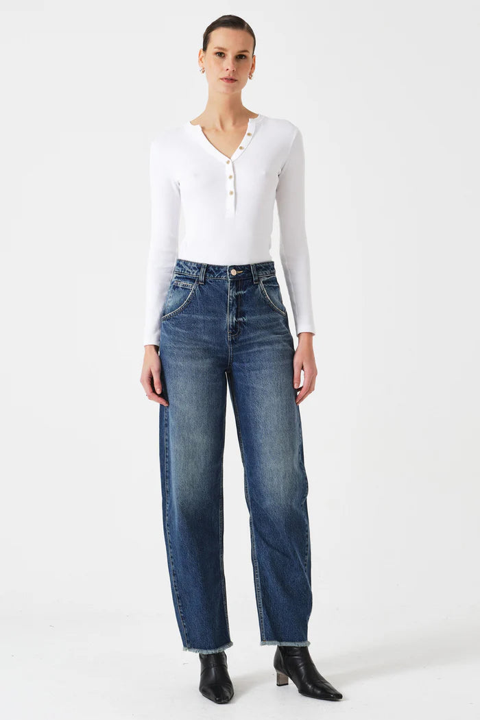 A person stands facing forward against a plain white background, wearing a white long-sleeve henley shirt, Seventy & Mochi Bo Barrel Leg Jeans - Phoenix in rigid blue denim, and black ankle boots.