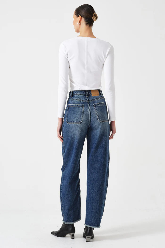 A person stands facing away in a white long-sleeve top, Seventy & Mochi's Bo Barrel Leg Jeans - Phoenix (super high-rise, back cutout, barrel leg), and black ankle boots against a plain background.