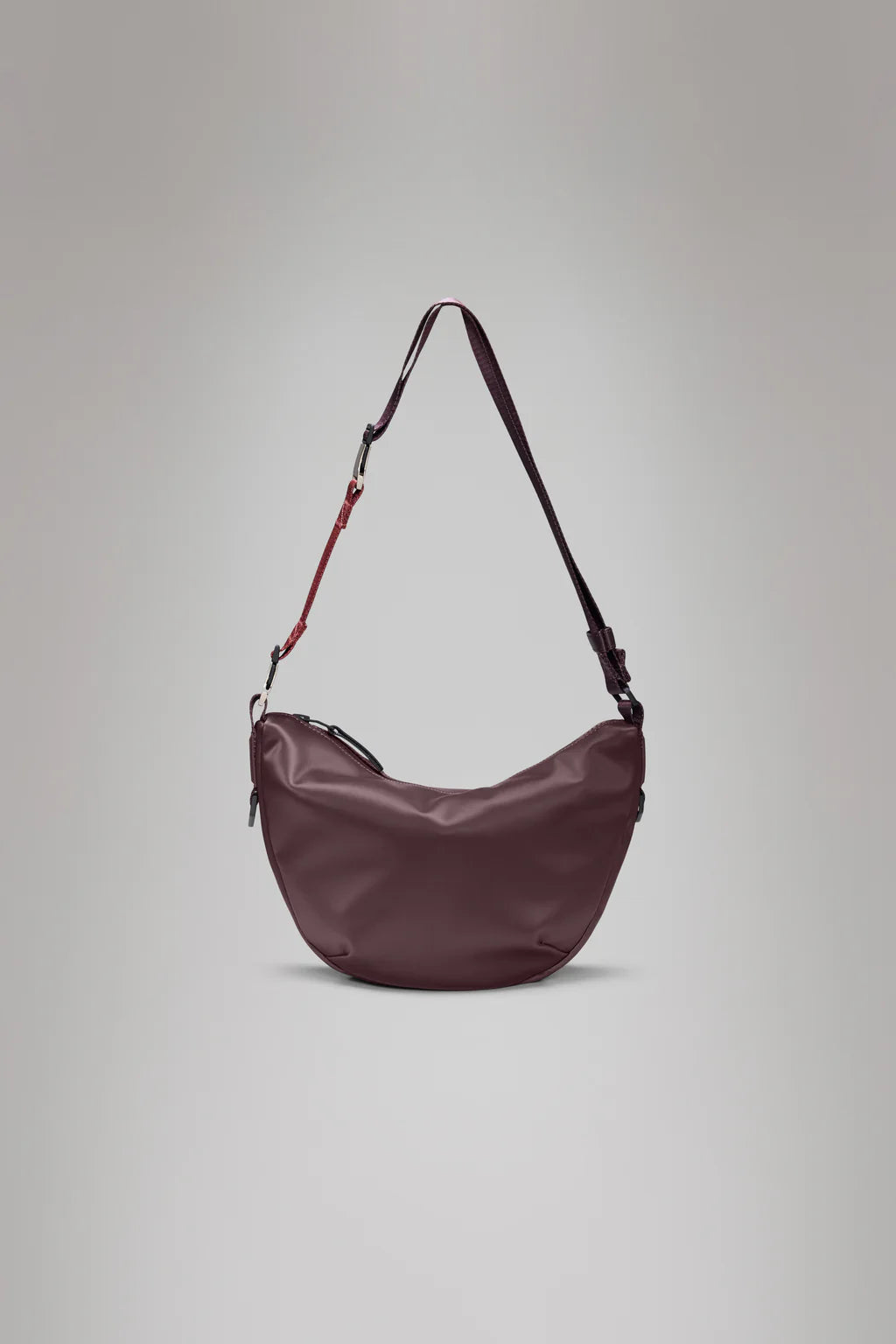 The Rains Valera Shoulder Bag, crescent-shaped with an adjustable strap in maroon, is showcased on a plain gray background. This chic waterproof bag is perfect for everyday use, combining style and practicality.