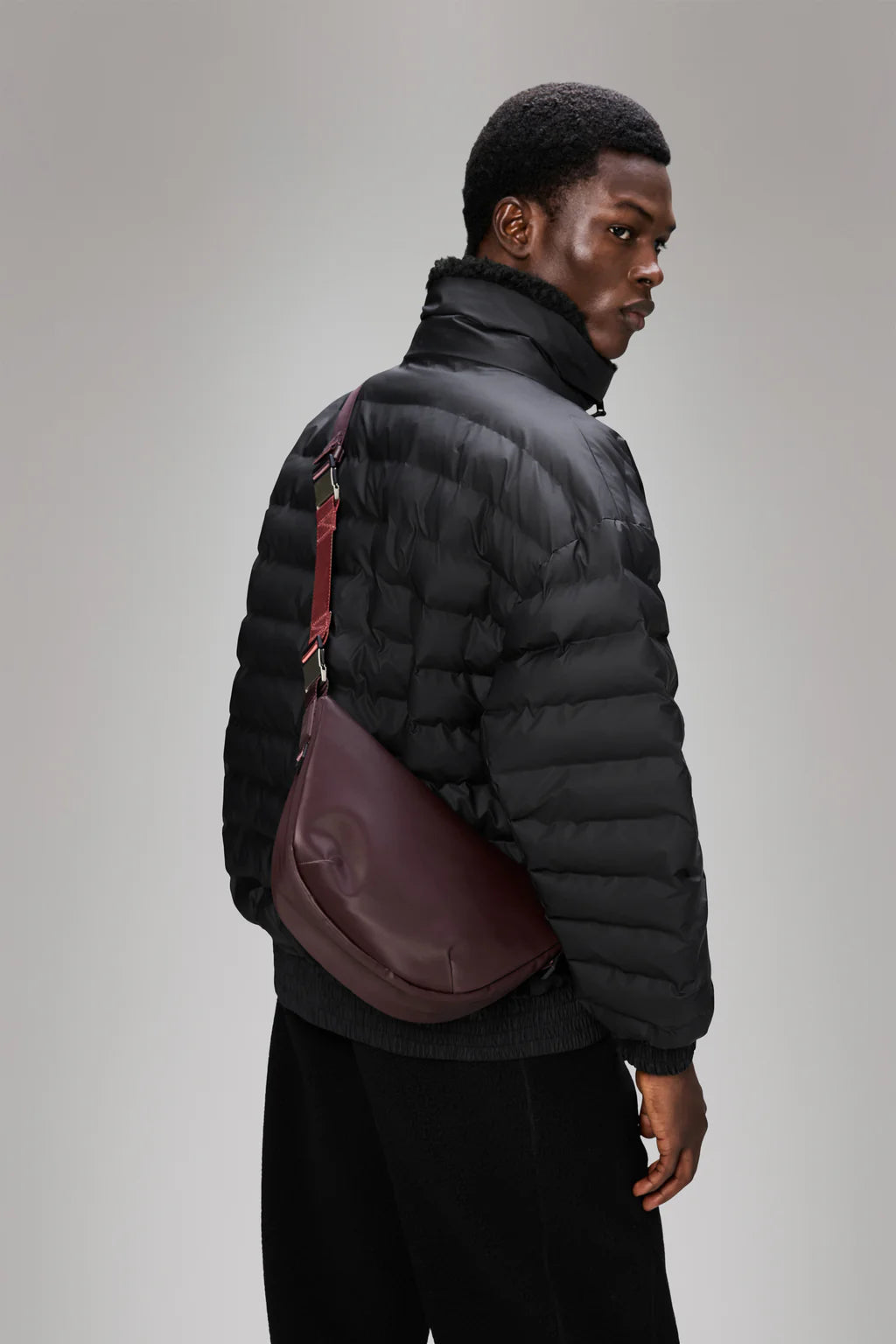 A man in a black puffer jacket and dark pants carries the Rains Valera Shoulder Bag, standing against a plain gray background while looking over his shoulder.