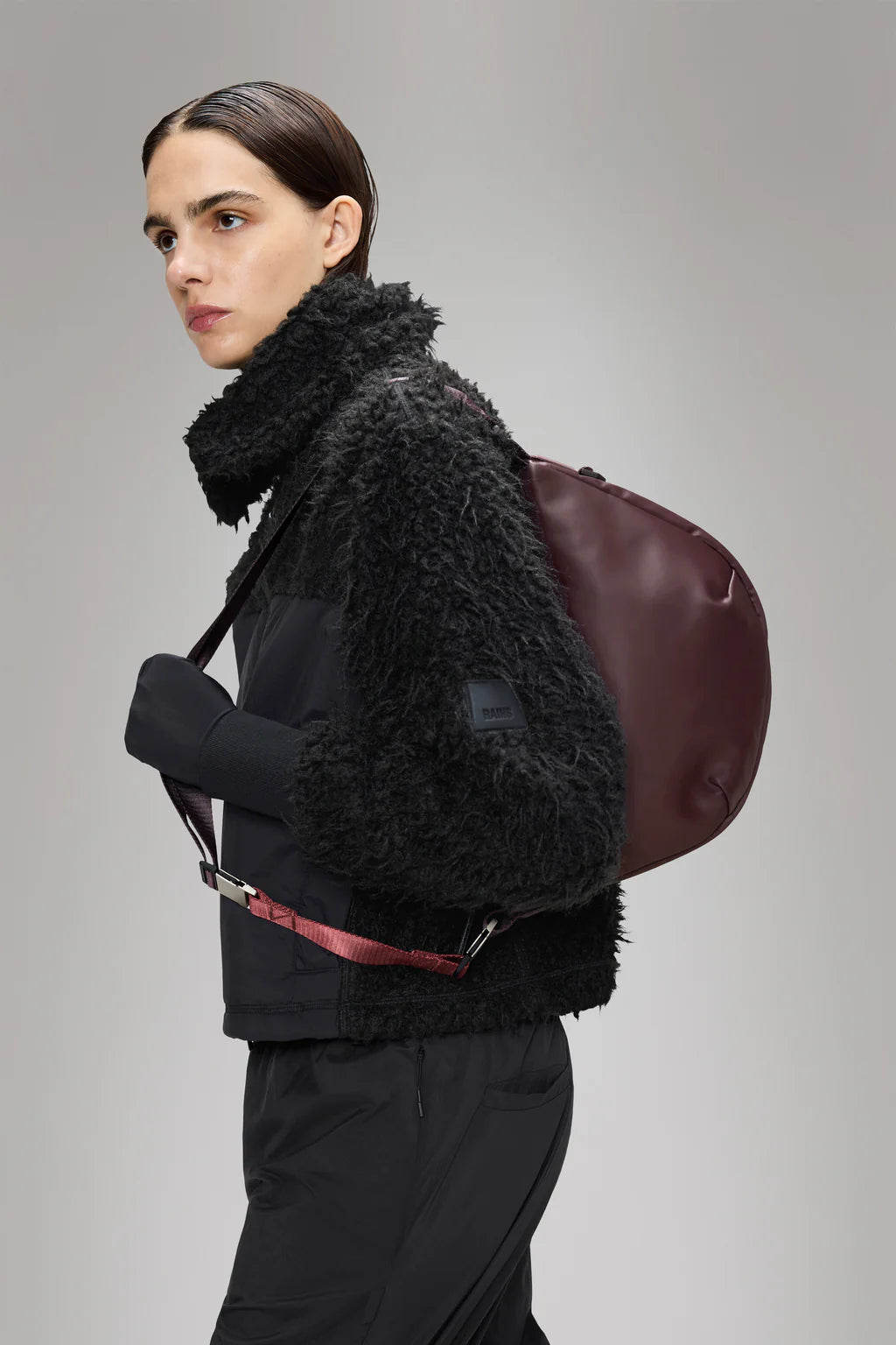 A person in a black textured jacket and pants carries the Rains Valera Shoulder Bag over one shoulder, standing against a plain grey background.