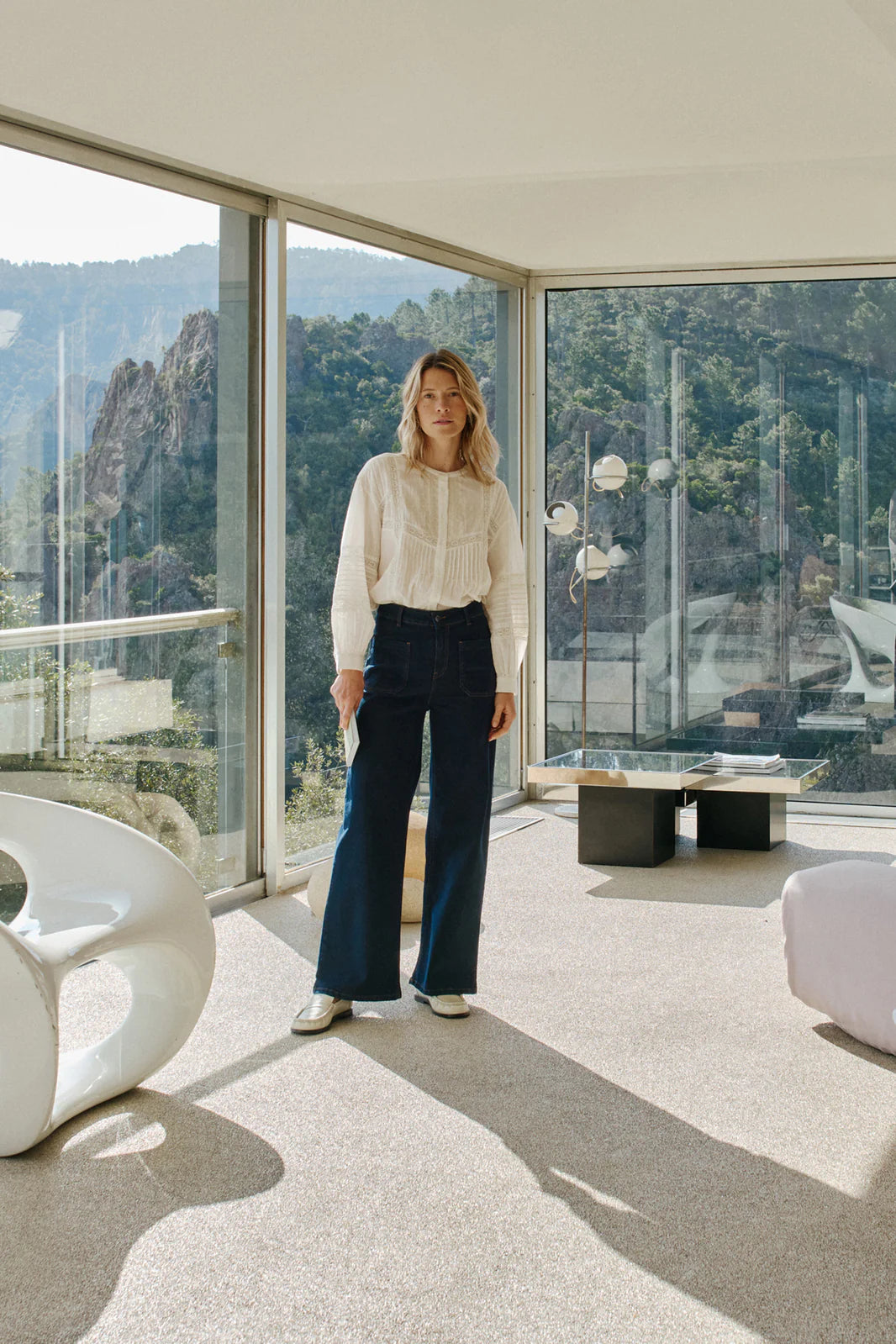 Indoors by large glass windows with mountain views, a person wears a white blouse and Indi & Cold’s Wide Leg Denim Trousers - Navy in a modern, minimal living space.