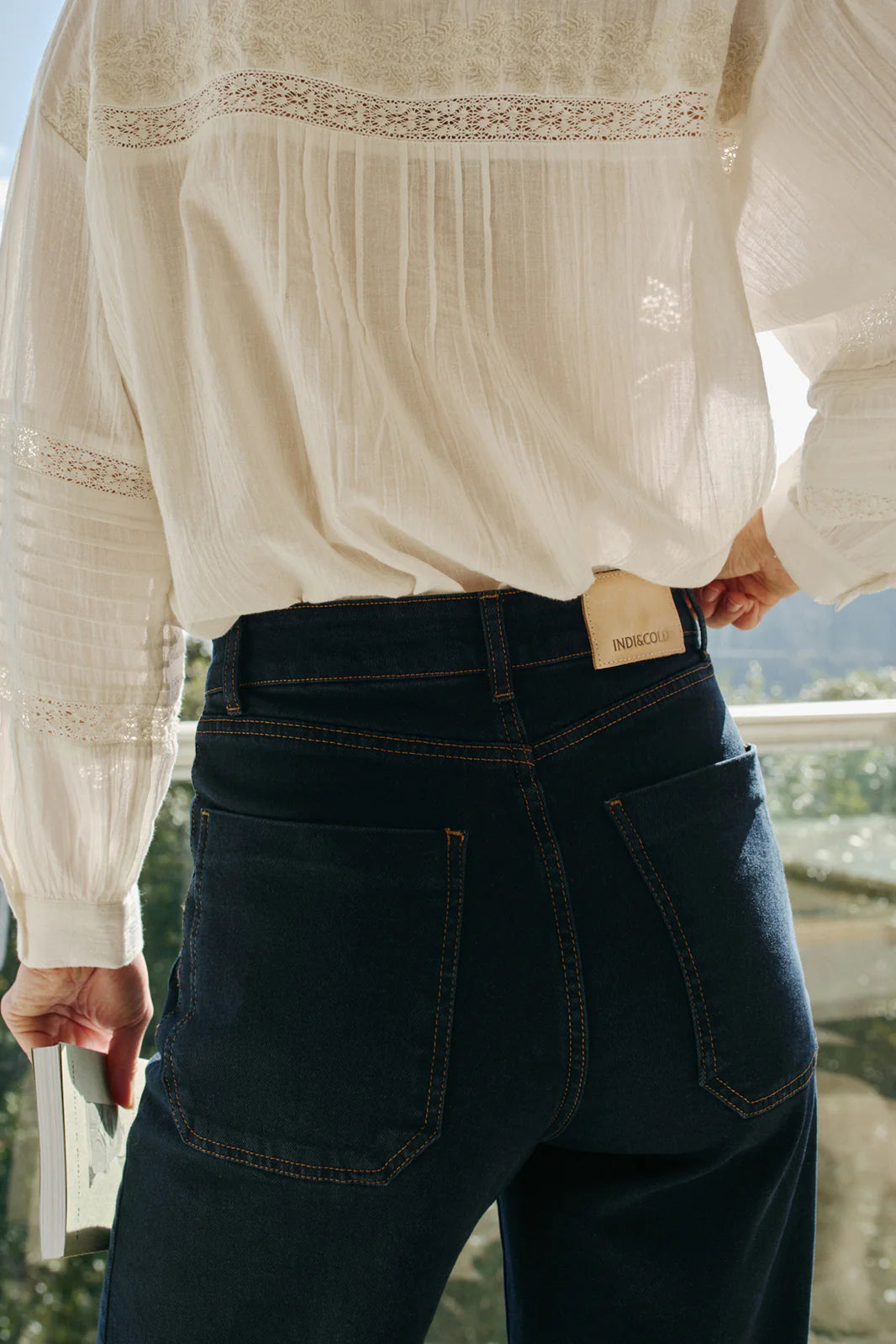 A person wearing a white long-sleeve blouse and Indi & Cold's Wide Leg Denim Trousers - Navy, holding a book outdoors with greenery in the background.