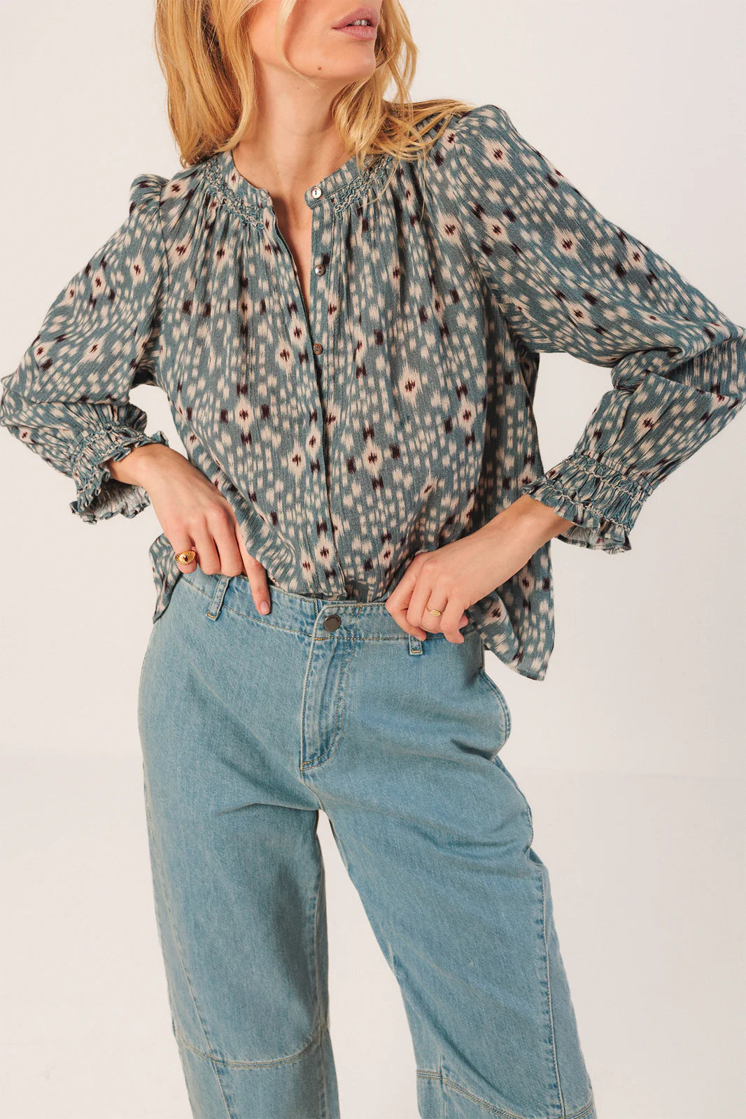 A woman poses in the Indi & Cold Smock Detail Shirt in Vintage Blue with ruffled cuffs, paired with light blue high-waisted jeans, against a plain background.