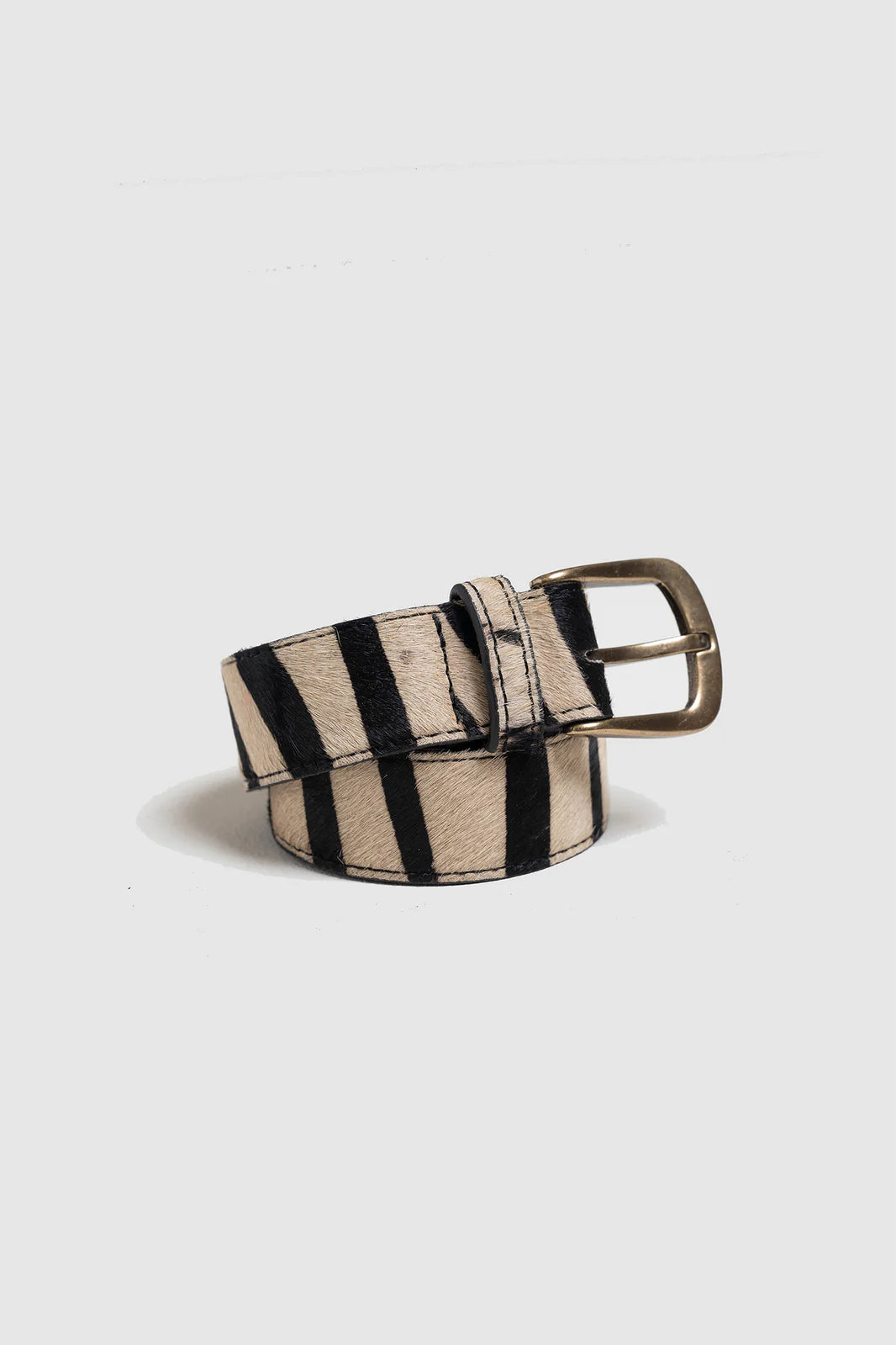 The Indi & Cold Horsehide Belt - Black features a rolled-up animal-print leather design with black and beige zebra stripes and a brass buckle, displayed against a plain light background.