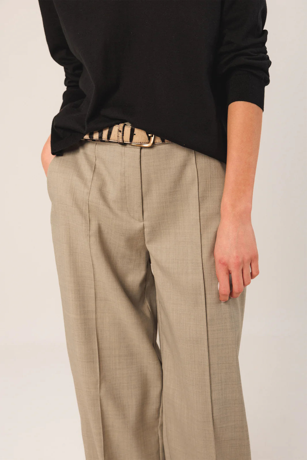 A person is seen from torso to upper legs, wearing a black long-sleeve top, beige pleated trousers, and the Indi & Cold Horsehide Belt - Black.