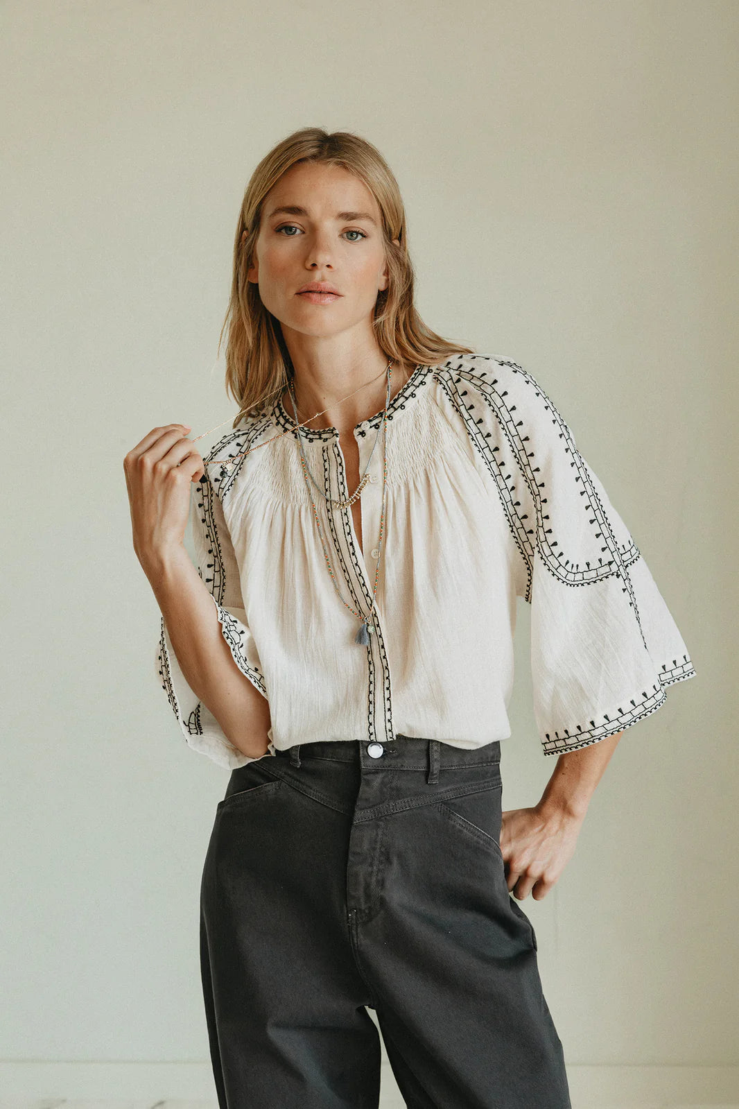 A woman stands indoors wearing the Indi & Cold Embroidered Blouse in off white, featuring black embroidery and wide sleeves, made from organic cotton, paired with high-waisted black pants.