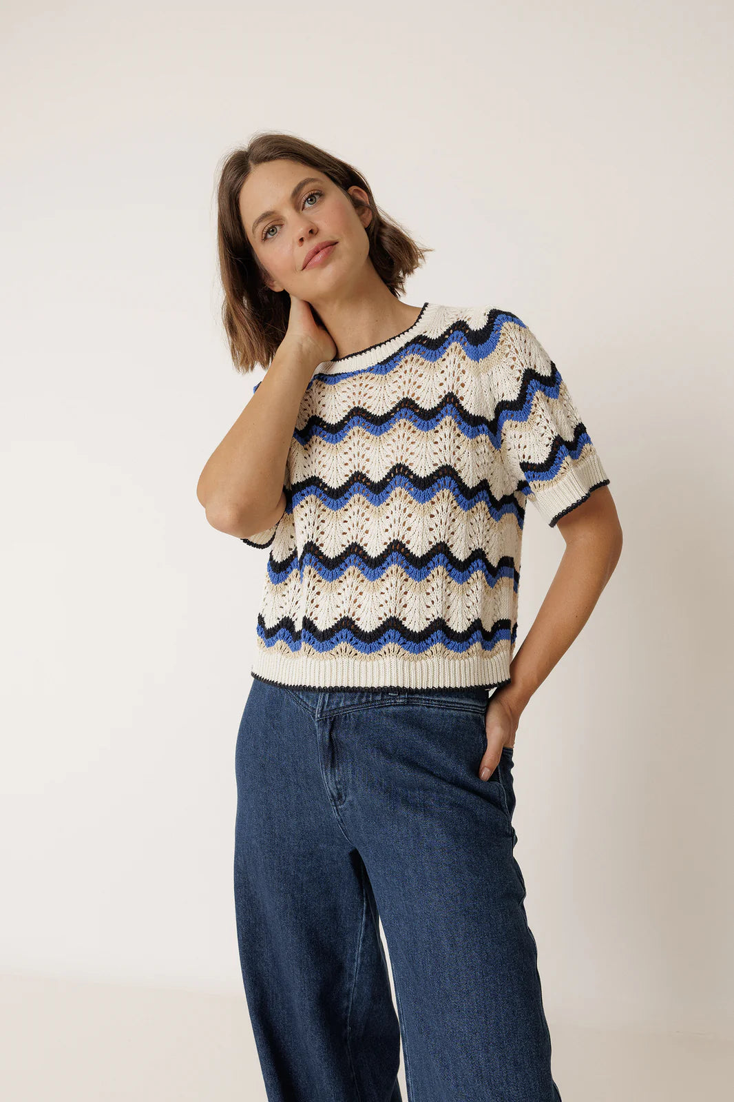 A woman wears the Indi & Cold Wavy Crochet Sweater in blue—a short-sleeve white sweater with a blue and black zig-zag pattern—paired with blue jeans, standing against a plain background.