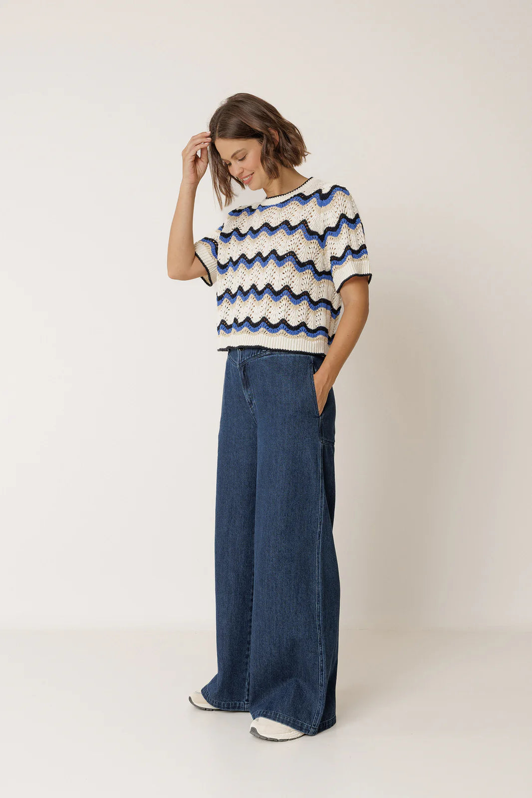 A woman stands against a plain background wearing the Indi & Cold Wavy Crochet Sweater in blue, styled with wide-leg blue jeans and white sneakers.
