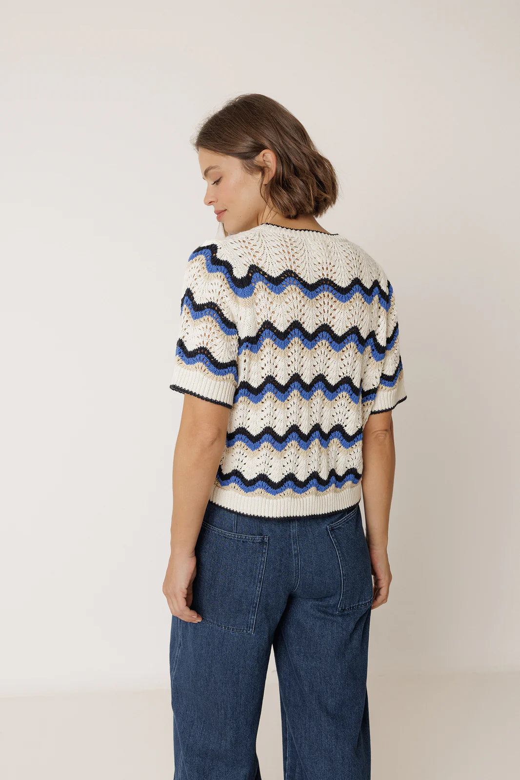 A woman with short brown hair stands with her back to the camera, wearing the Indi & Cold Wavy Crochet Sweater in blue and blue jeans.
