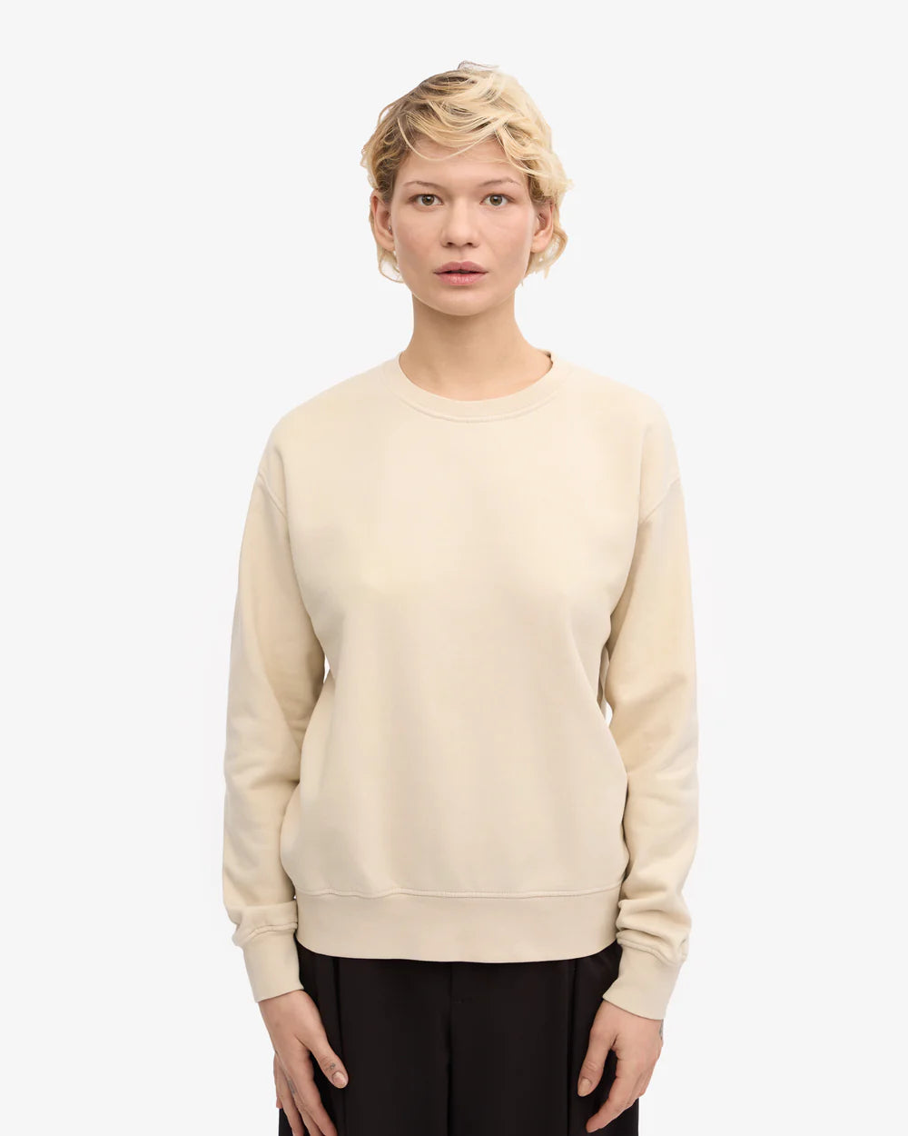 A person with short, wavy blonde hair stands facing forward against a white background, wearing Colorful Standard's Classic Organic Crew in dense knit beige cotton and black pants.