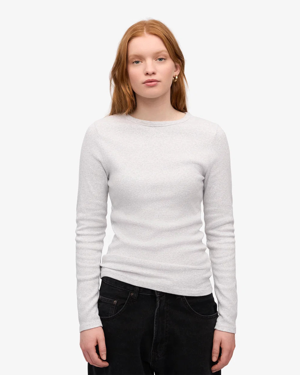 A person with long red hair is standing against a plain background, wearing the Colorful Standard Women Organic Rib LS T-Shirt in Snow Melange and black jeans.
