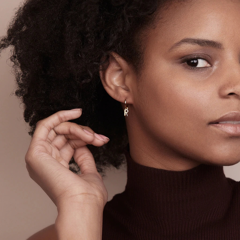 A black woman wearing a turtle neck and gold hoop earrings embraces the spirit of the Art Deco era with her stylish outfit. Her alphabet initials, RJL, add a personal touch to her Rachel Jackson London Art Deco Initial Hoop - Gold.