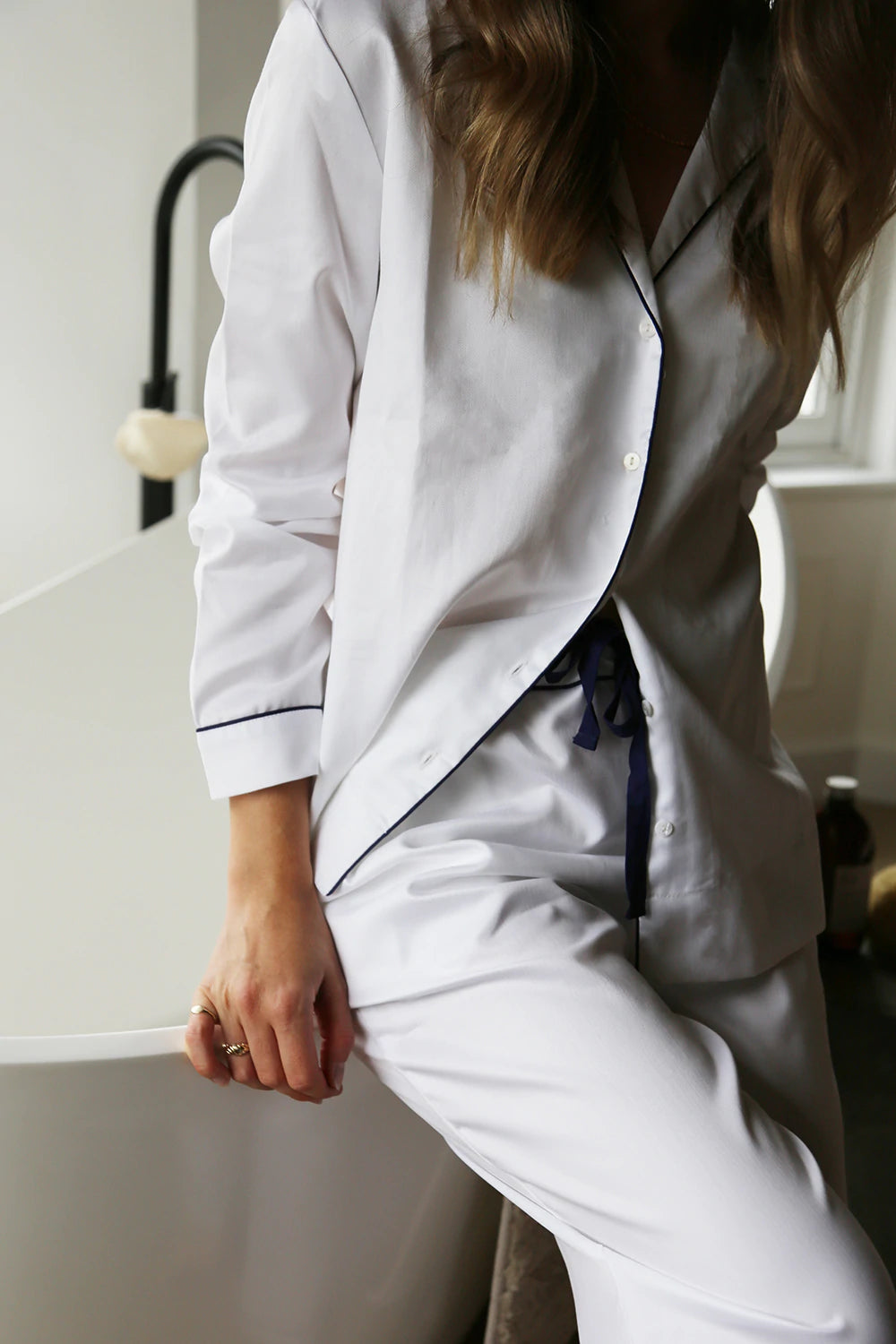 A woman in BREATHE organic cotton pyjamas, specifically the Herringbone White organic cotton pyjama set, leaning against a bathtub.