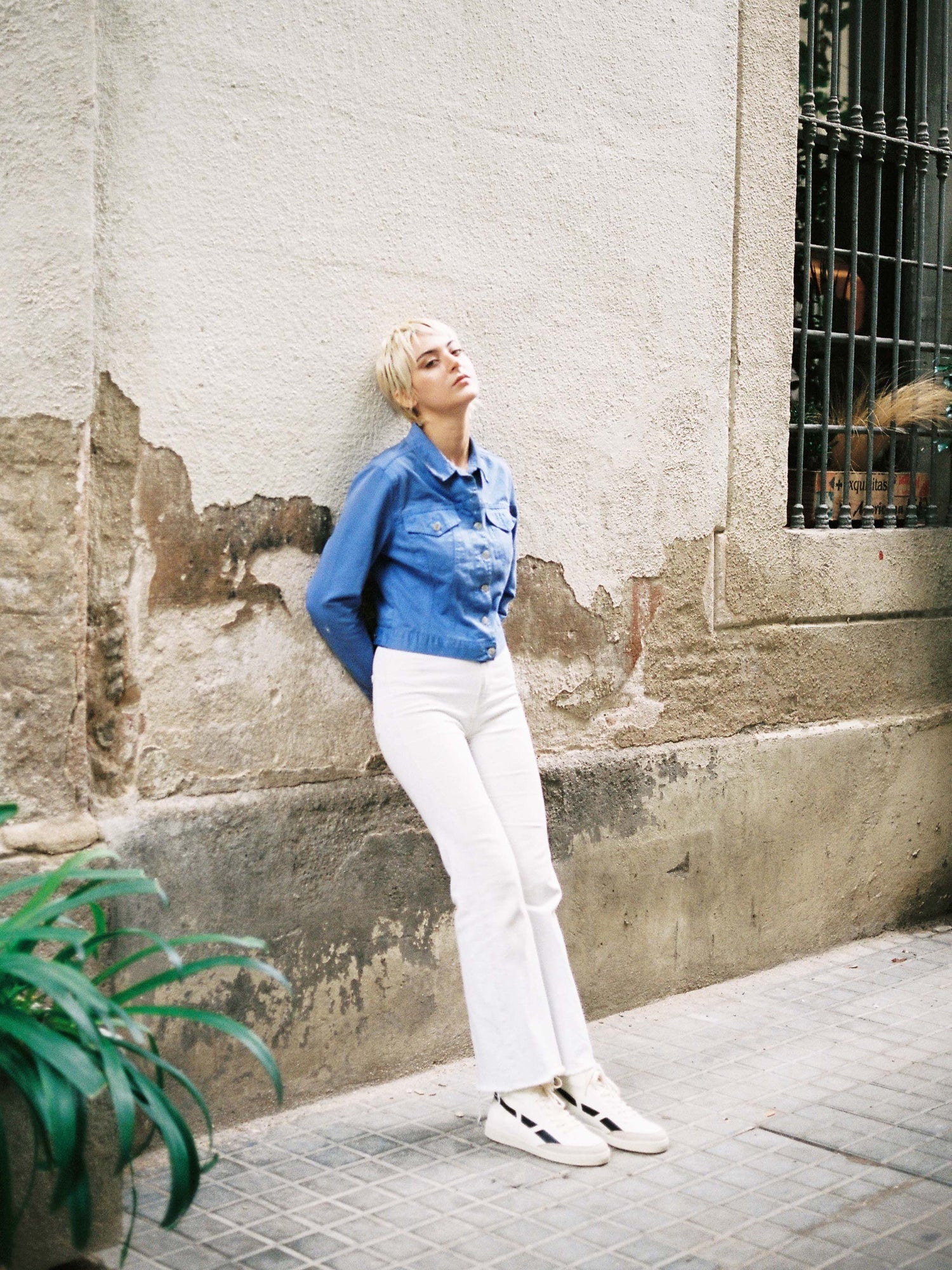A woman leaning against a wall wearing white pants and a blue shirt made from SAYE's Modelo '89 Hi Sneakers - Black.