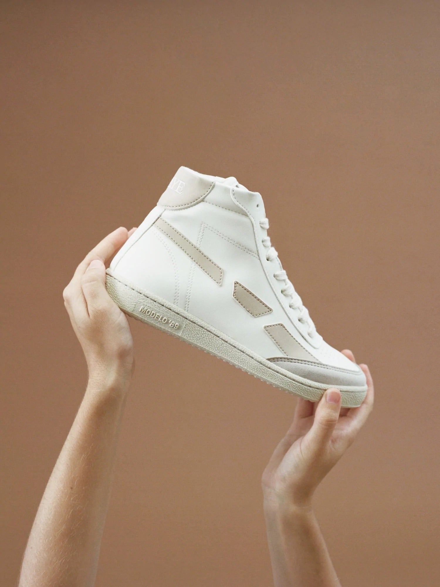 A pair of hands holding up a SAYE Modelo '89 Hi Sneaker - Beige made from bio-based vegan leather.