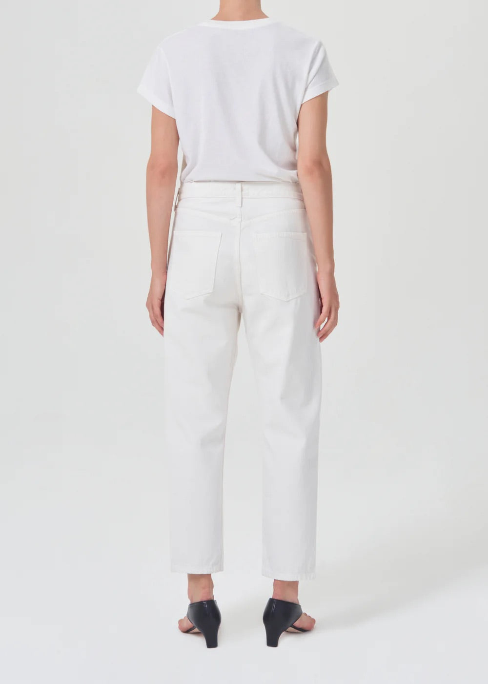 person wears white straight leg agolde jeans with black sandals, highlighting the back of the jeans against  a white studio backdrop