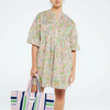 person wears the Dover beach dress whic hhas an all over paisley print in light greens, pinks and blues. The person is holding a large striped shopper bag and is in a white studio. 