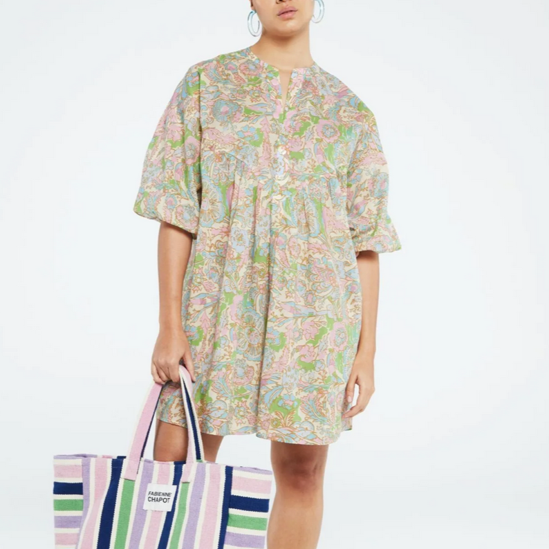 person wears the Dover beach dress whic hhas an all over paisley print in light greens, pinks and blues. The person is sitting on a wicker style seat in a white studio. 