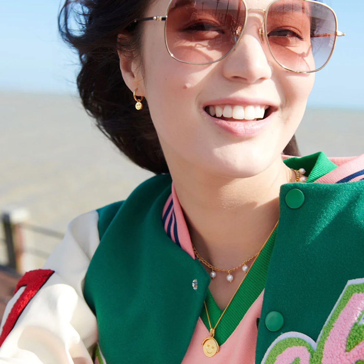 A Rachel Jackson woman, wearing sunglasses and a baseball jacket with retro star detail, adorned with the Happy Face Huggie Hoop Earrings - Gold.