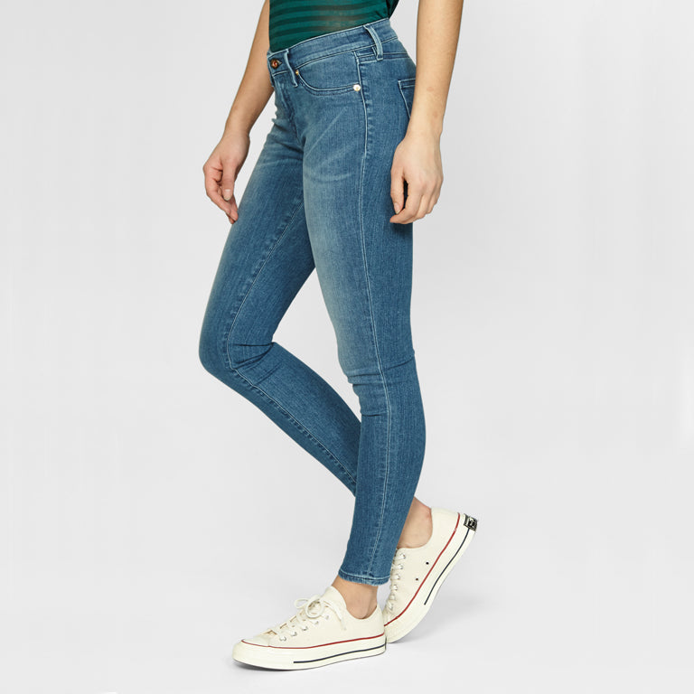 A woman donning a comfortable and stretchy SPRAY Skinny - Golden Rivet Prosecco jean in a mid-blue hue, paired with a stylish green top from Denham.