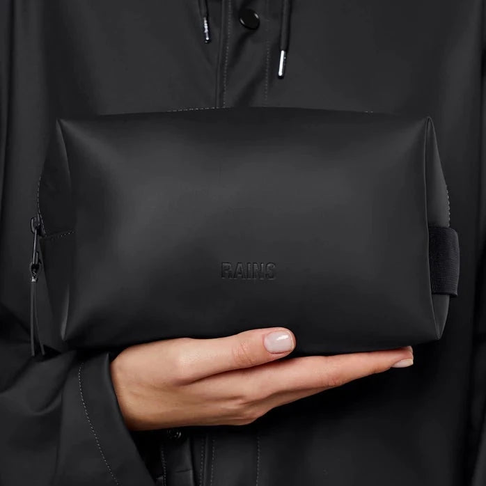 person wearing a black jacket is holding small matte black washbag with zip closure and webbed side handle 