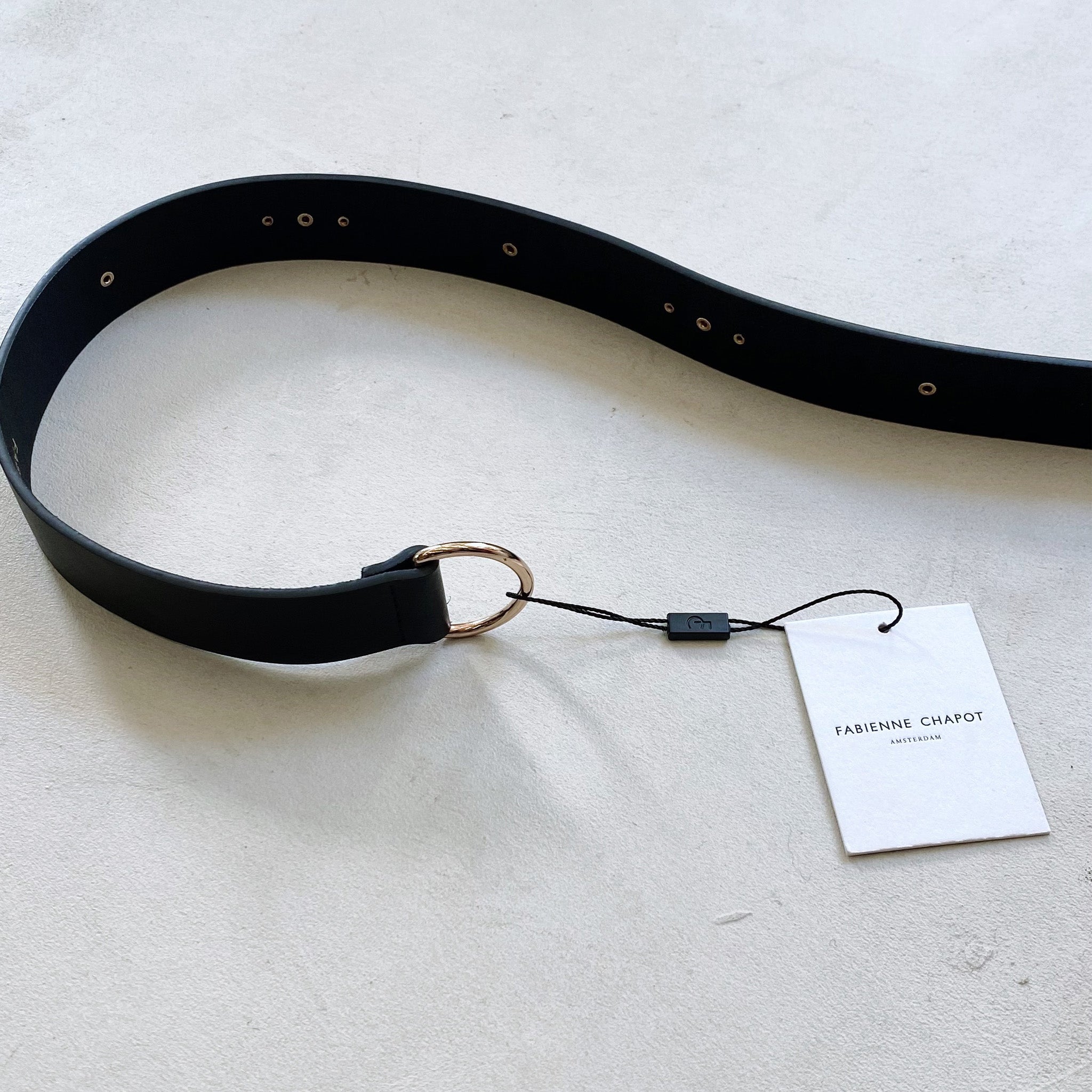 A Fabienne Chapot black leather belt with a tag on it, featuring gold studs.