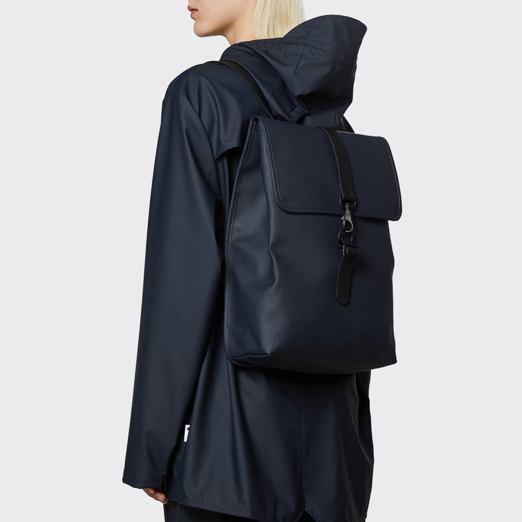 person wears navy rains coat and Matte navy rucksack with flap and black rubber central hook fastening