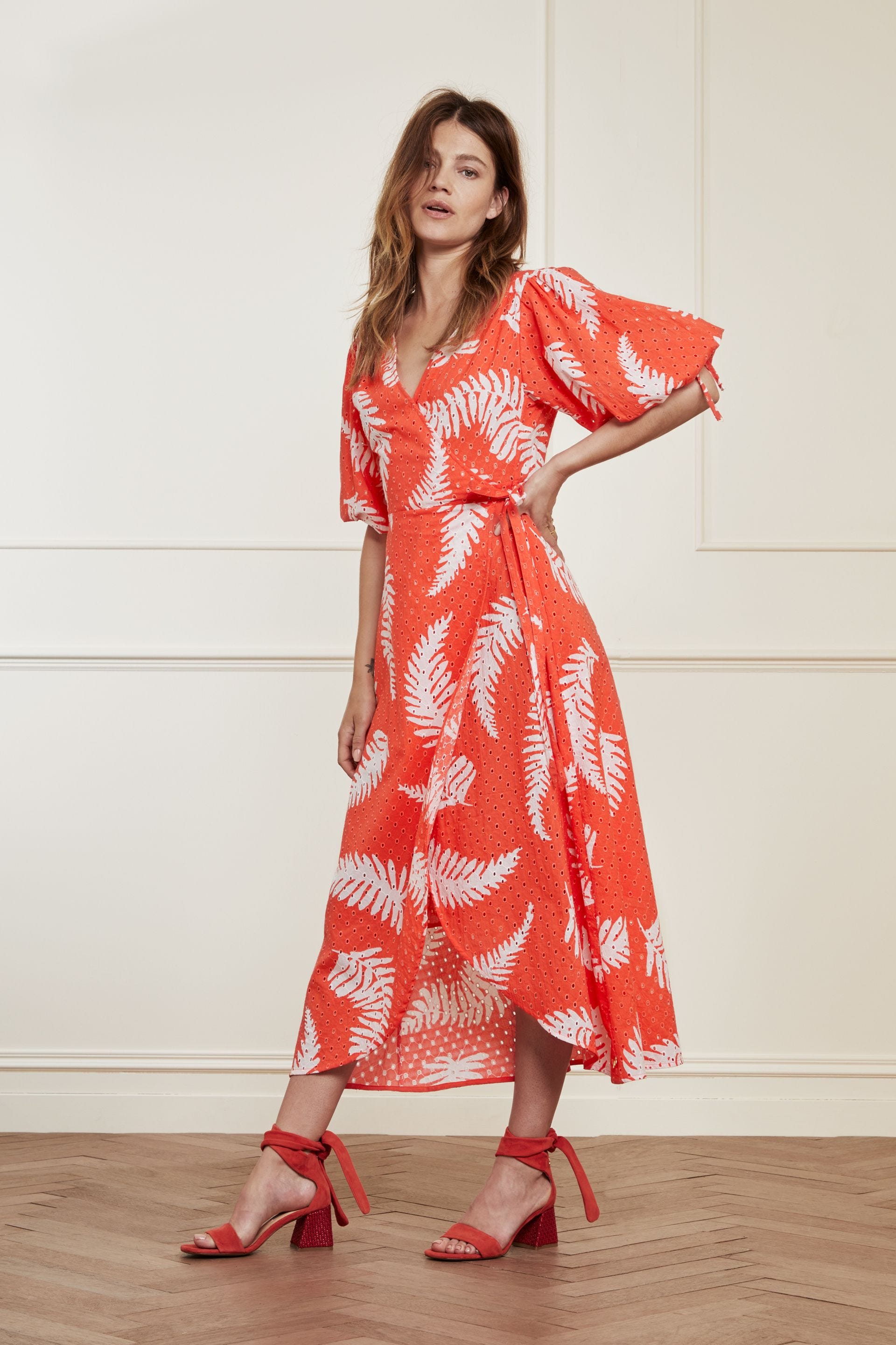 A model wearing the Fabienne Chapot Charlie Broderie Dress in Hot Coral, made of 100% GOTS-certified cotton, featuring a v-neckline and falling to midi length.